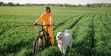 a lady in sunglasses and an orange tracksuit sitting on a bike in a grassy field with a retriever next to her