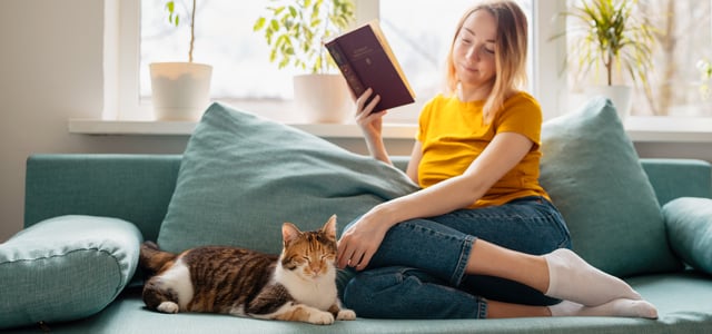 A woman sitting on a sofa reading a book next to her cat.