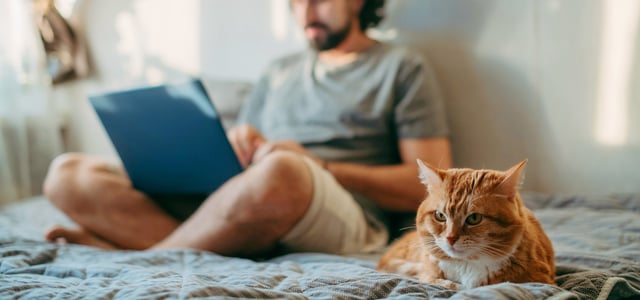 A man sat on a bed crossed legged with his ginger cat.