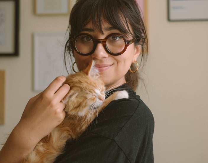 A woman with dark hair and glasses sitting on a couch holding an orange kitten.