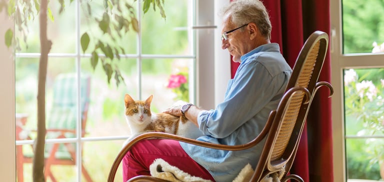 A cat sat on the lap of an elderly man in a rattan chair.