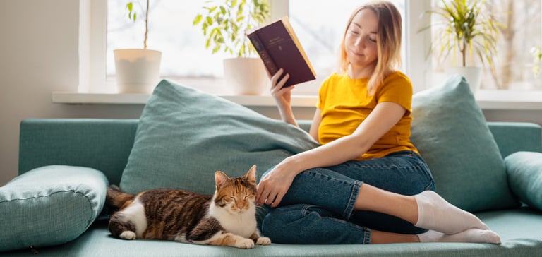 A woman sitting on a sofa reading a book next to her cat.
