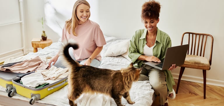 Two women sitting on a bed with a suitcase and a cat.