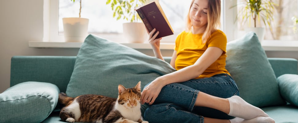 A woman sitting on a sofa reading a book next to her cat.