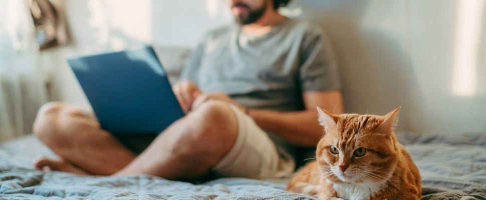 A man sat on a bed crossed legged with his ginger cat.