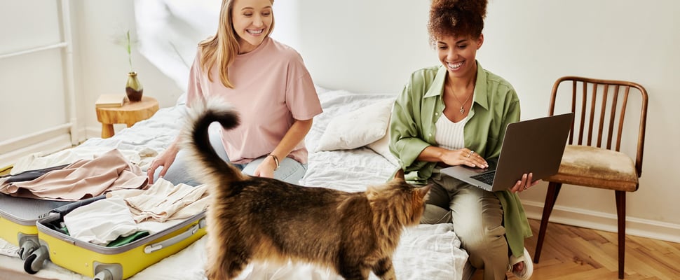 Two women sitting on a bed with a suitcase and a cat.